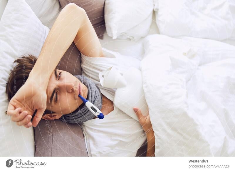 Kranker junger Mann mit Thermometer im Mund, der zu Hause auf dem Bett liegt Farbaufnahme Farbe Farbfoto Farbphoto Deutschland Innenaufnahme Innenaufnahmen
