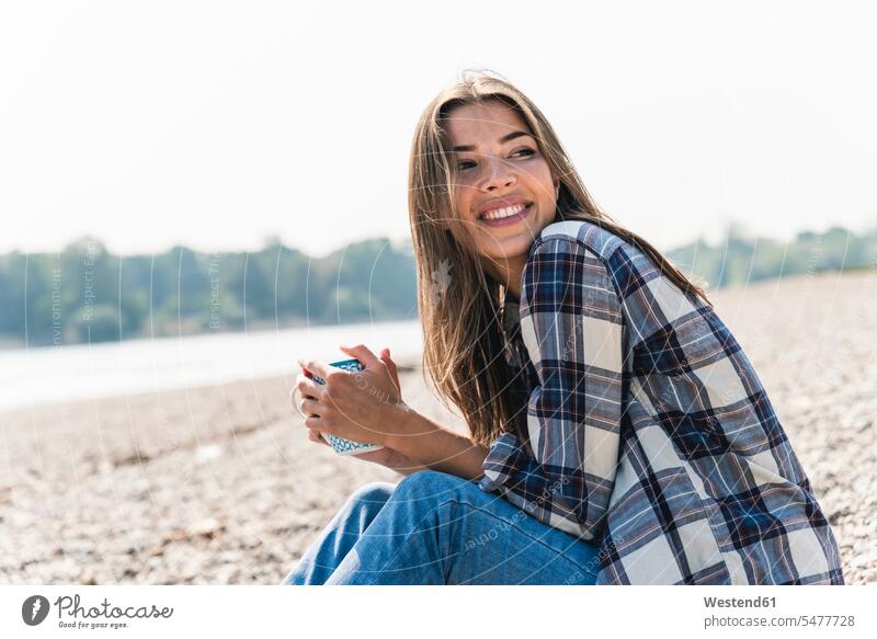Glückliche junge Frau mit einem Becher in der Hand am Flussufer sitzend glücklich glücklich sein glücklichsein weiblich Frauen sitzt halten Ufer Erwachsener
