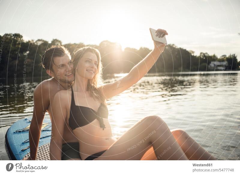 Glückliches Paar sitzt auf einem Paddelbrett auf einem See und macht Smartphone-Selfies Badebekleidung Bikinis Telekommunikation telefonieren Handies Handys
