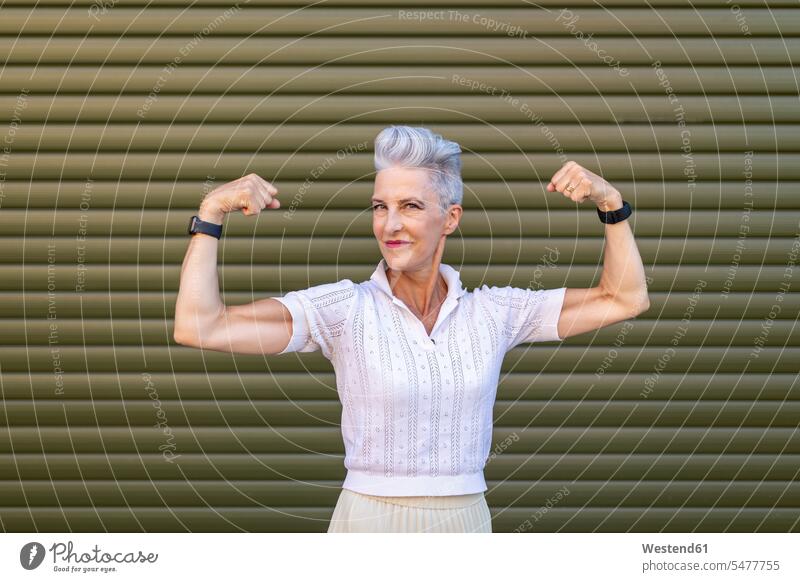 Ältere Frau, die Muskeln anspannt, während sie gegen den Fensterladen steht Farbaufnahme Farbe Farbfoto Farbphoto Außenaufnahme außen draußen im Freien Tag