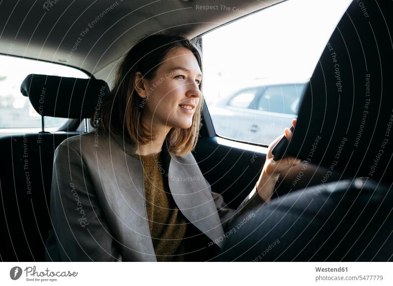 Lächelnde Frau sitzt auf dem Rücksitz eines Autos und hält ein Mobiltelefon weiblich Frauen sitzen sitzend lächeln halten Ruecksitz Ruecksitze Rücksitze Handy