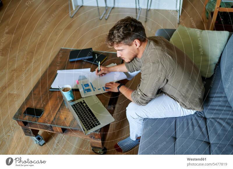 Männlicher Unternehmer schreibt auf Papier, während er zu Hause mit seinem Laptop sitzt Farbaufnahme Farbe Farbfoto Farbphoto Innenaufnahme Innenaufnahmen innen
