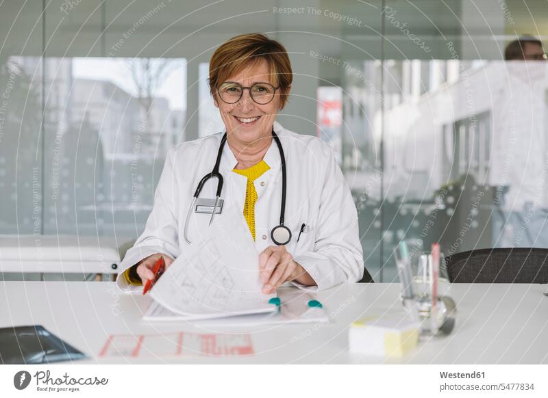 Porträt eines lächelnden Arztes am Schreibtisch sitzend Job Berufe Berufstätigkeit Beschäftigung Jobs Dokumente Papiere Unterlagen Glasscheiben Gesundheit