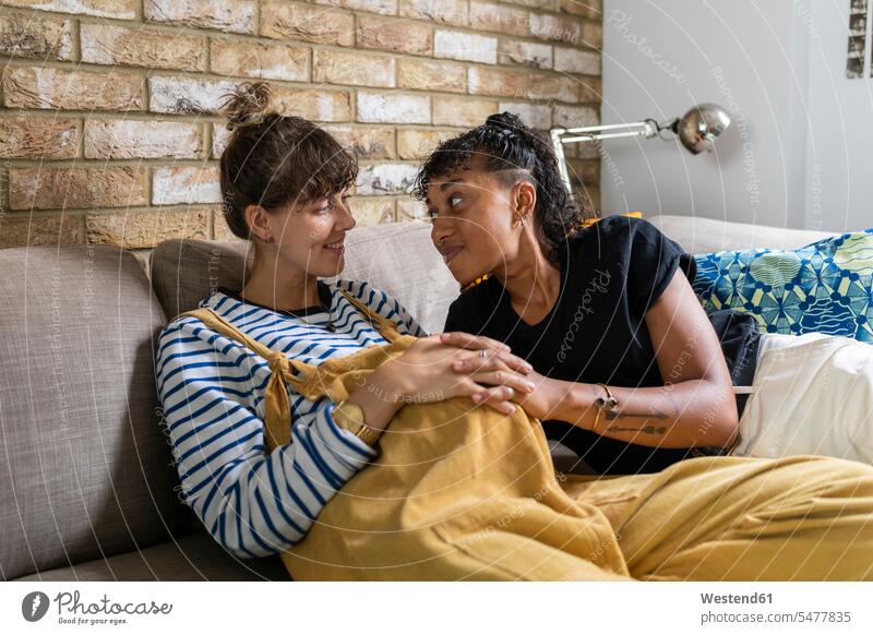 Lächelndes lesbisches Paar sitzt mit gefalteten Händen zu Hause auf dem Sofa Farbaufnahme Farbe Farbfoto Farbphoto Innenaufnahme Innenaufnahmen innen drinnen