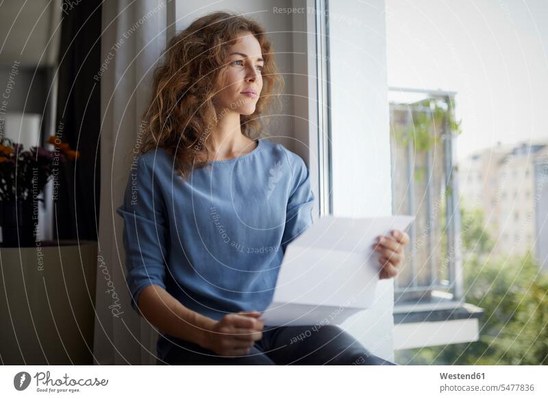 Frau hält Papier in der Hand, während sie zu Hause auf dem Fensterbrett sitzt Farbaufnahme Farbe Farbfoto Farbphoto Innenaufnahme Innenaufnahmen innen drinnen