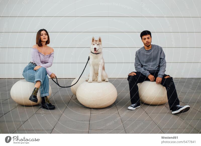 Freund und Freundin sitzen von Hund mit sozialen Abstand auf weißen Beton Ball gegen die Wand Farbaufnahme Farbe Farbfoto Farbphoto Außenaufnahme außen draußen