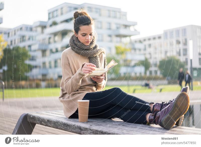 Frau mit Kaffee zum Mitnehmen sitzt im Herbst auf einer Bank und liest ein Buch lesen Lektüre weiblich Frauen Bücher sitzen sitzend Sitzbänke Bänke Sitzbank