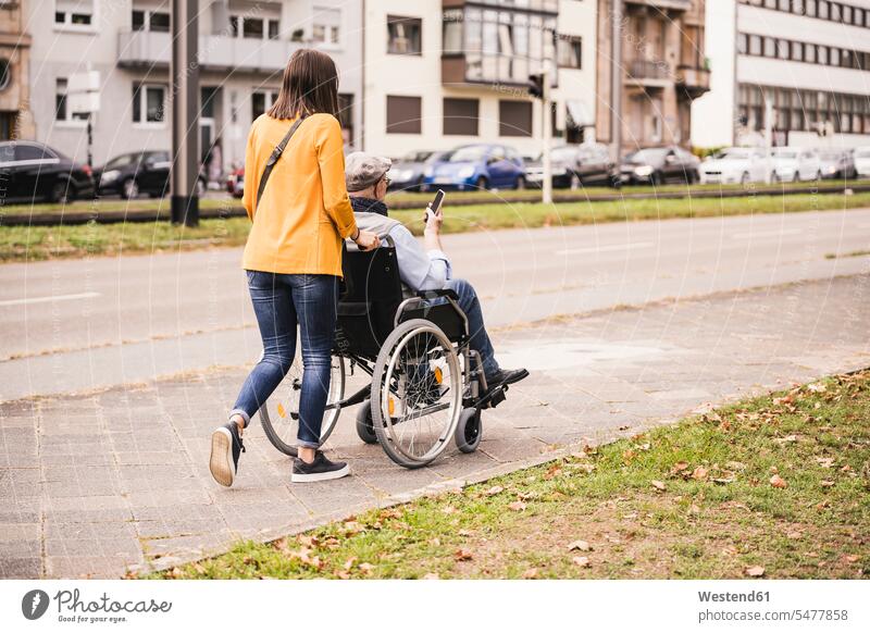 Rückenansicht einer jungen Frau, die einen älteren Mann mit einem Smartphone im Rollstuhl auf dem Bürgersteig schiebt Leute Menschen People Person Personen