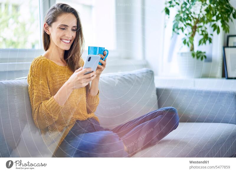 Lächelnde junge Frau auf Sofa sitzend mit Smartphone und Tasse Kaffee Leute Menschen People Person Personen Alleinstehende Alleinstehender Singles