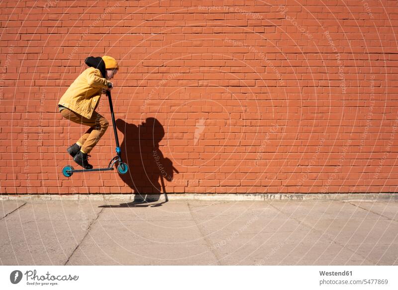 Junge führt bei sonnigem Wetter einen Stunt mit einem Schubsroller auf dem Bürgersteig gegen eine Ziegelmauer aus Farbaufnahme Farbe Farbfoto Farbphoto