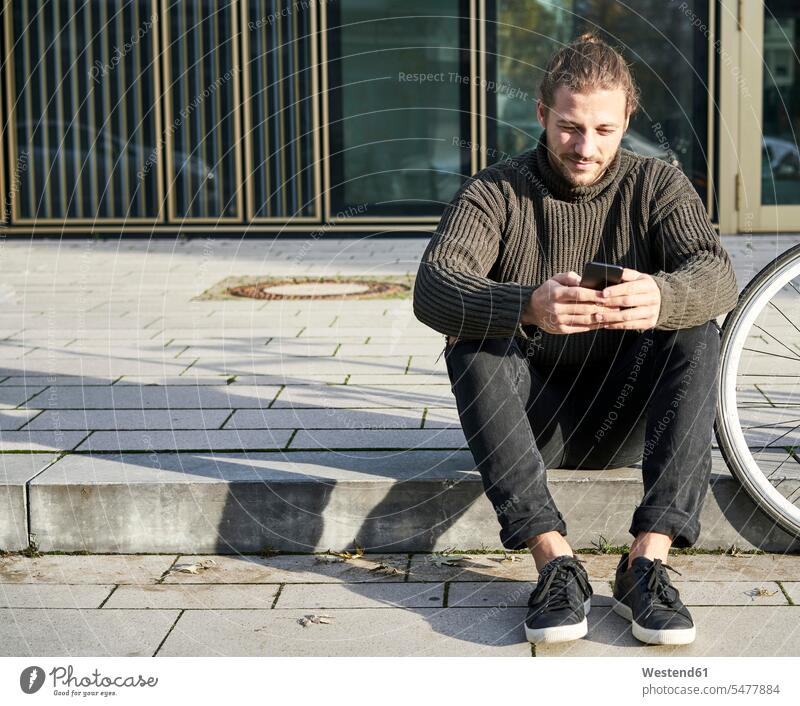 Porträt eines bärtigen jungen Mannes, der auf einem Bordstein neben einem Fahrrad sitzt und ein Mobiltelefon benutzt daneben Männer männlich Bordsteinkante