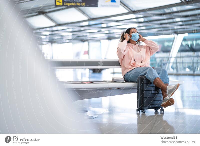 Junge Frau hört Musik, während sie auf dem Flughafen sitzend eine schützende Gesichtsmaske trägt Farbaufnahme Farbe Farbfoto Farbphoto Innenaufnahme