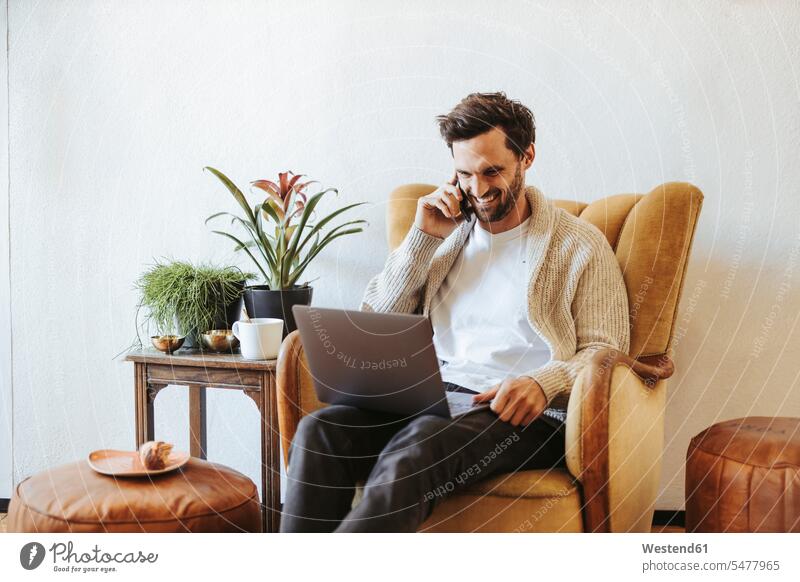 Lächelnder Mann am Telefon, der zu Hause auf einem Sessel sitzt und auf den Laptop schaut Leute Menschen People Person Personen Europäisch Kaukasier kaukasisch