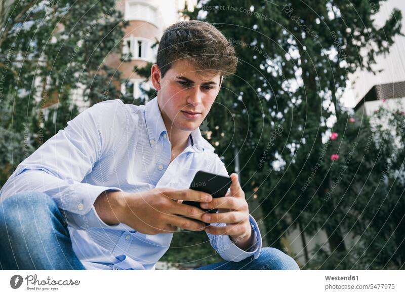 Hübscher junger Mann benutzt Smartphone, während er in der Stadt an Bäumen sitzt Farbaufnahme Farbe Farbfoto Farbphoto Spanien Außenaufnahme außen draußen