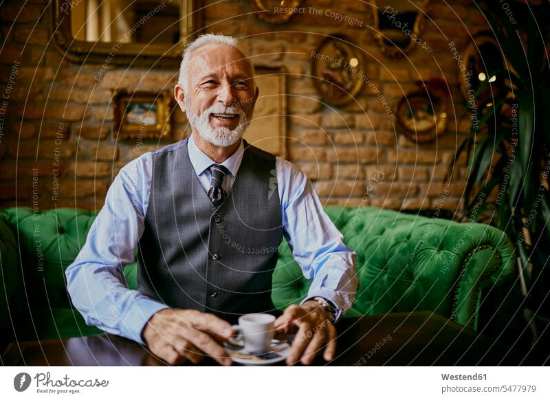 Porträt eines eleganten älteren Mannes, der lächelnd auf einer Couch in einem Café sitzt Portrait Porträts Portraits Cafe Kaffeehaus Bistro Cafes Cafés
