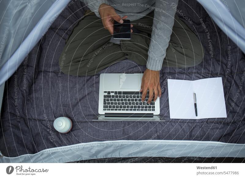 Mann sitzt im Zelt und benutzt Laptop und Mobiltelefon, Teilansicht Männer männlich Notebook Laptops Notebooks Zelte Smartphone iPhone Smartphones sitzen