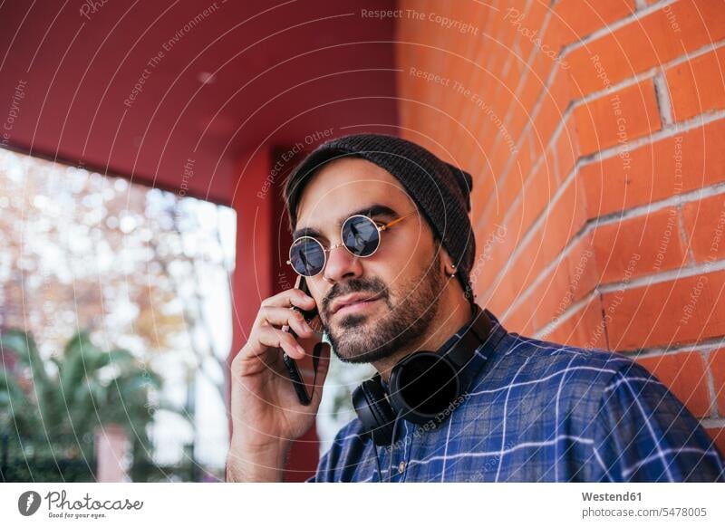 Modischer junger Mann mit Sonnenbrille beim Telefonieren gegen eine Backsteinmauer Farbaufnahme Farbe Farbfoto Farbphoto Außenaufnahme außen draußen im Freien