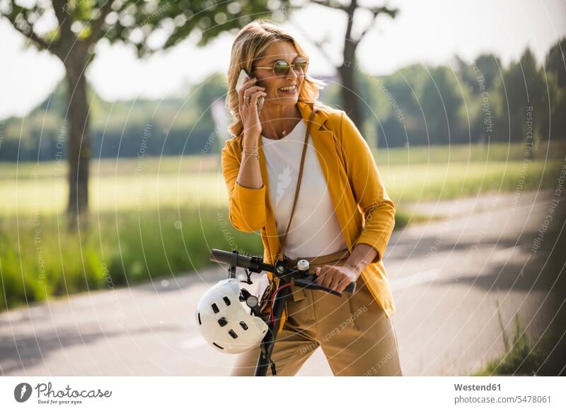 Glückliche reife Frau, die ein Smartphone benutzt, während sie mit einem Roller am Straßenrand steht Farbaufnahme Farbe Farbfoto Farbphoto Außenaufnahme außen