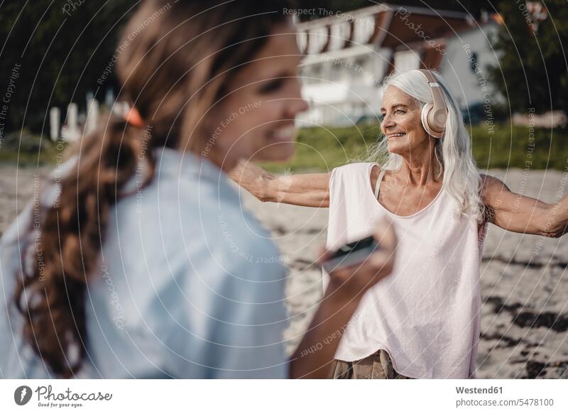 ältere Frau tanzt am Strand, Tochter lacht im Vordergrund Generation Kopfhoerer Telekommunikation telefonieren Handies Handys Mobiltelefon Mobiltelefone hoeren