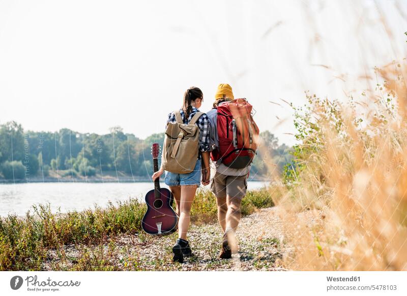 Rückansicht eines jungen Paares mit Rucksäcken und Gitarre beim Spaziergang am Flussufer Rucksack Gitarren gehen gehend geht Fluesse Fluß Flüsse Pärchen