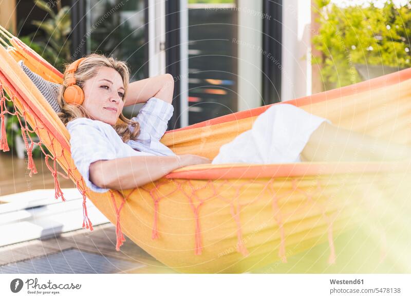 Porträt einer nachdenklichen, reifen Frau, die in der Hängematte auf der Terrasse liegt und mit Kopfhörern Musik hört Hängematten Kopfhoerer entspannen relaxen