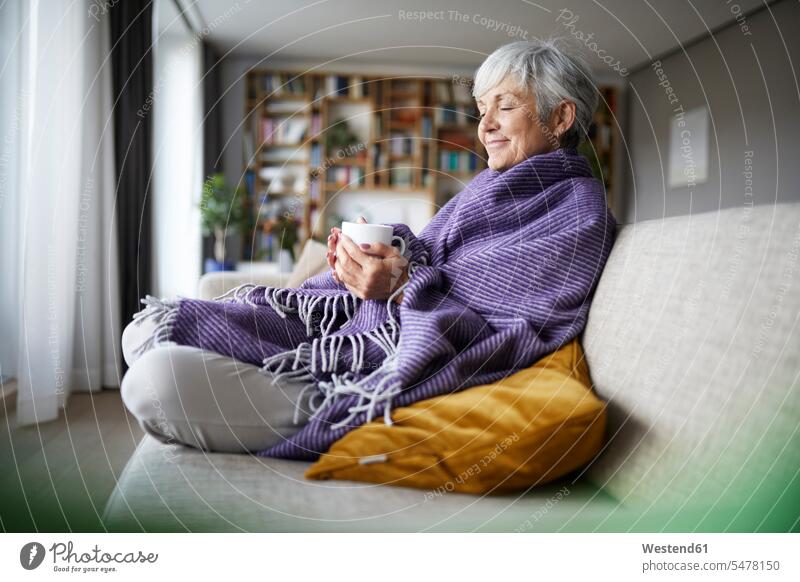 Nachdenkliche ältere Frau mit Decke, die zu Hause auf dem Sofa sitzend eine Kaffeetasse hält Farbaufnahme Farbe Farbfoto Farbphoto Innenaufnahme Innenaufnahmen
