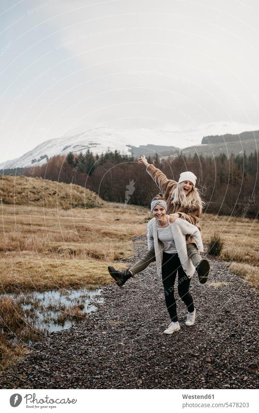 Großbritannien, Schottland, glückliche junge Frau trägt Freund Huckepack in ländlicher Landschaft Freundinnen Glück glücklich sein glücklichsein Portrait