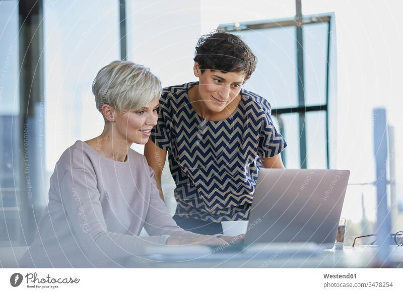 Zwei lächelnde Geschäftsfrauen teilen Laptop am Schreibtisch im Büro Teilen Sharing Office Büros arbeiten Arbeit Kollegin Kolleginnen Businesswomen