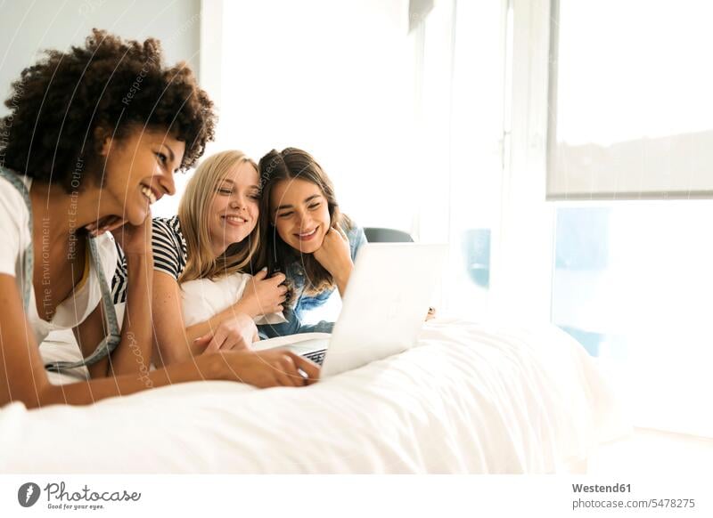 Drei glückliche Freundinnen liegen auf dem Bett und teilen sich einen Laptop liegend liegt Betten Notebook Laptops Notebooks Glück glücklich sein glücklichsein