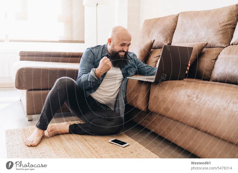 Glücklicher Geschäftsmann jubelt mit geballter Faust, während er im Wohnzimmer auf den Laptop schaut Farbaufnahme Farbe Farbfoto Farbphoto Freizeitkleidung