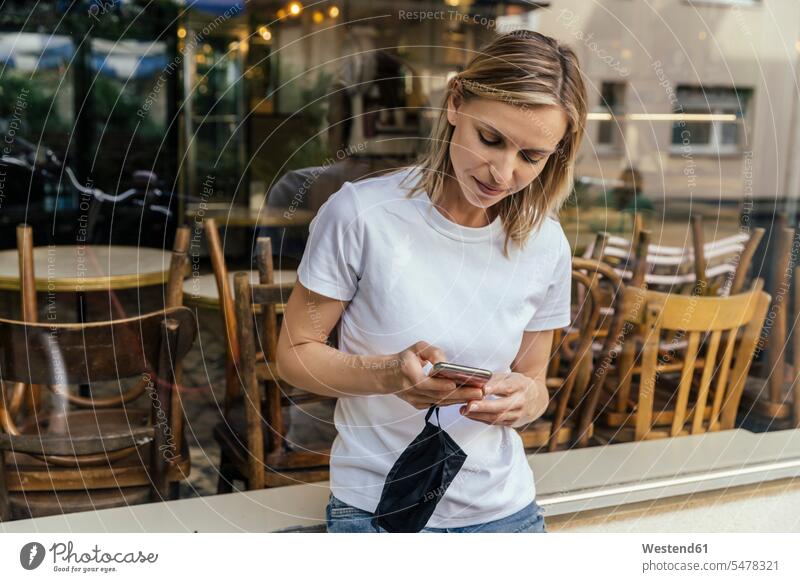 Porträt einer Frau mit Schutzmaske in der Hand Textnachrichten vor einem geschlossenen Café Leute Menschen People Person Personen Europäisch Kaukasier