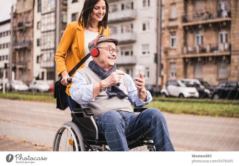 Lächelnde junge Frau schiebt glücklichen älteren Mann mit Kopfhörern und Smartphone im Rollstuhl Leute Menschen People Person Personen Europäisch Kaukasier