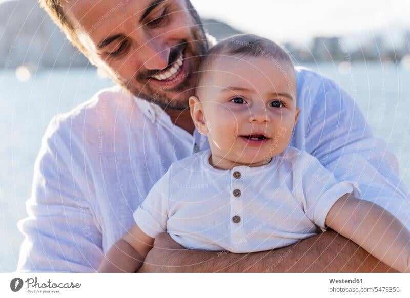 Nahaufnahme eines lächelnden Vaters, der seinen süßen Sohn bei Sonnenuntergang am Strand trägt Farbaufnahme Farbe Farbfoto Farbphoto Spanien