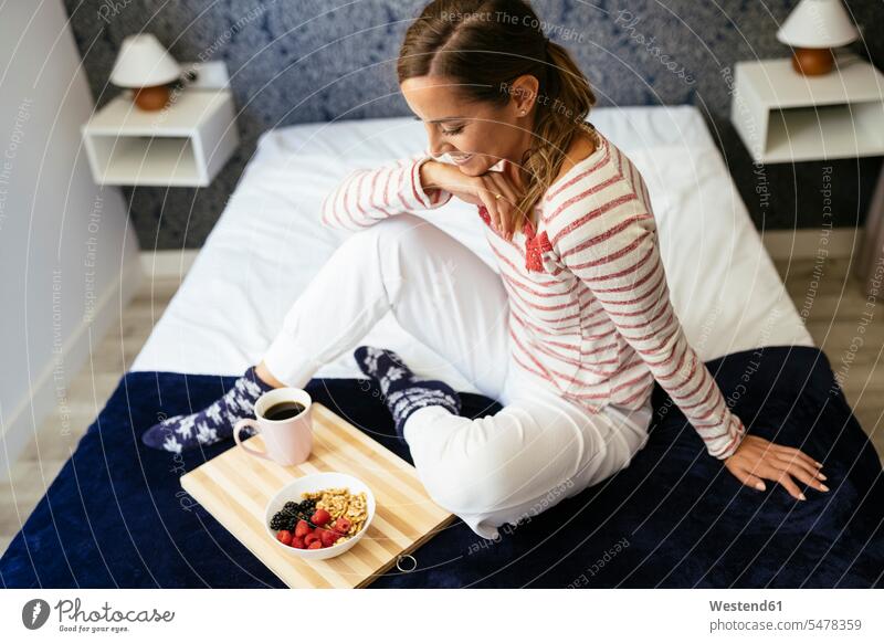 Lächelnde Frau betrachtet Essen und Kaffee, während sie zu Hause auf dem Bett sitzt Farbaufnahme Farbe Farbfoto Farbphoto Innenaufnahme Innenaufnahmen innen