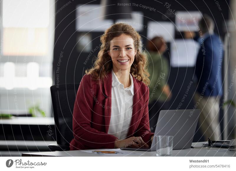 Portrait einer lächelnden Geschäftsfrau mit Laptop am Schreibtisch im Büro mit Kollegen im Hintergrund Arbeitskollege Arbeitskollegen Job Berufe Berufstätigkeit