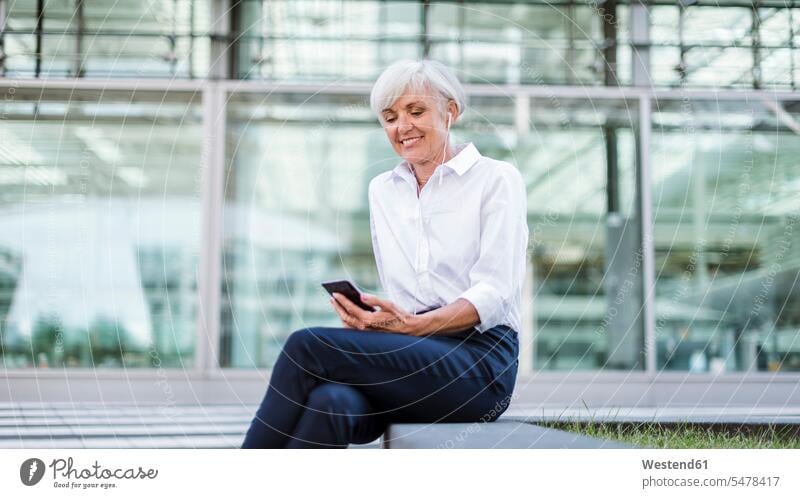 Ältere Geschäftsfrau sitzt draußen mit Smartphone und Ohrstöpseln Geschäftsfrauen Businesswomen Businessfrauen Businesswoman sitzen sitzend Frau weiblich Frauen