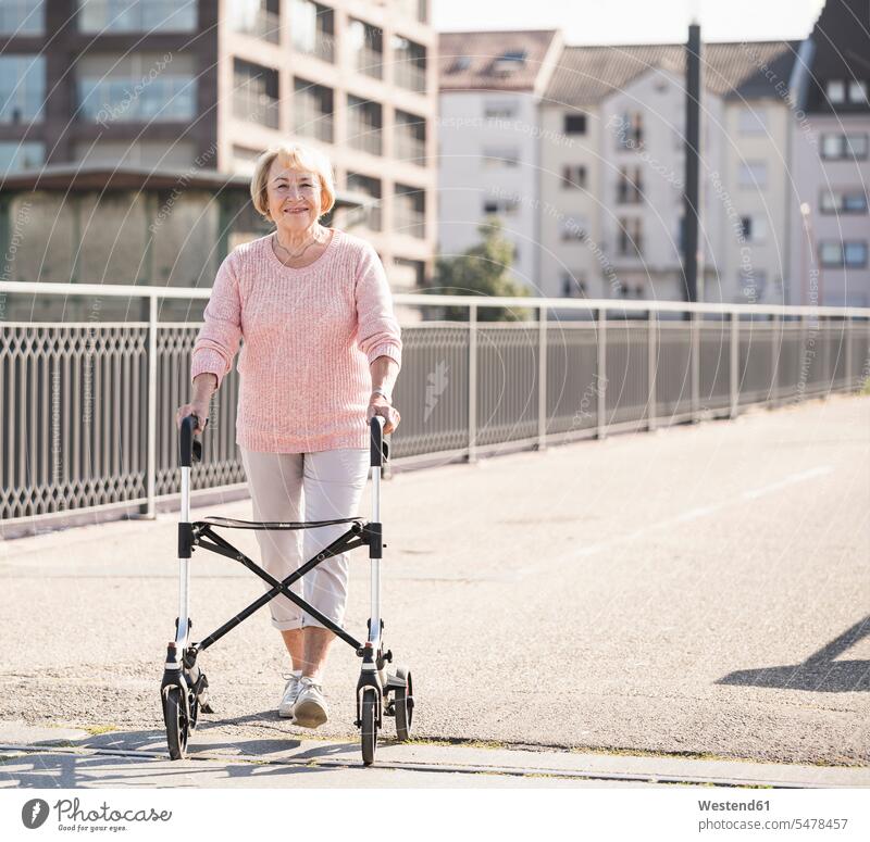 Ältere Frau mit Gehhilfe auf Rädern auf Fußgängerbrücke gehend geht Farben Farbtoene Farbton Farbtöne rosafarben gesellschaftliche Themen Soziales Thema