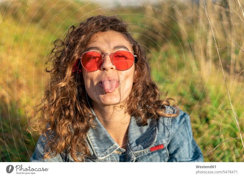 Porträt einer jungen Frau mit lockigem braunem Haar und roter Sonnenbrille, die die Zunge herausstreckt Sonnenbrillen Brille weiblich Frauen herausstrecken