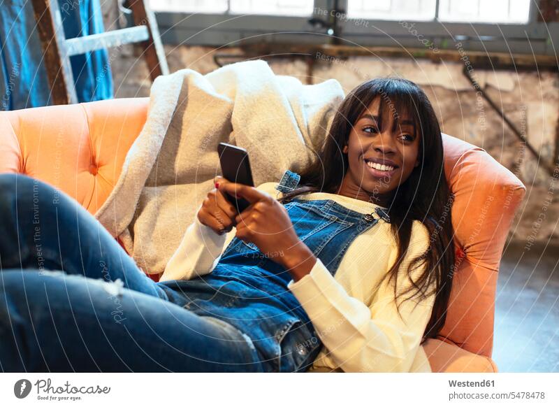Porträt einer lächelnden Frau, die zu Hause auf der Couch liegt und ihr Handy benutzt Smartphone iPhone Smartphones liegen liegend Loft Lofts Sofa Couches Liege