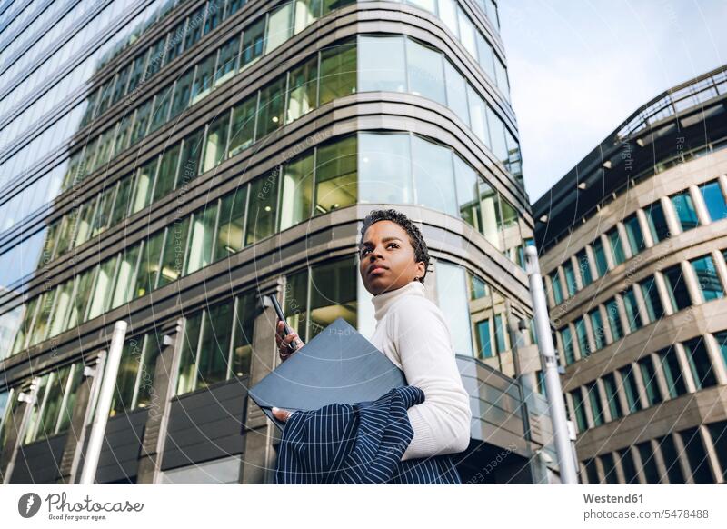 Geschäftsfrau schaut weg, während sie Handy und Laptop gegen moderne Gebäude in der Stadt hält Farbaufnahme Farbe Farbfoto Farbphoto Außenaufnahme außen draußen