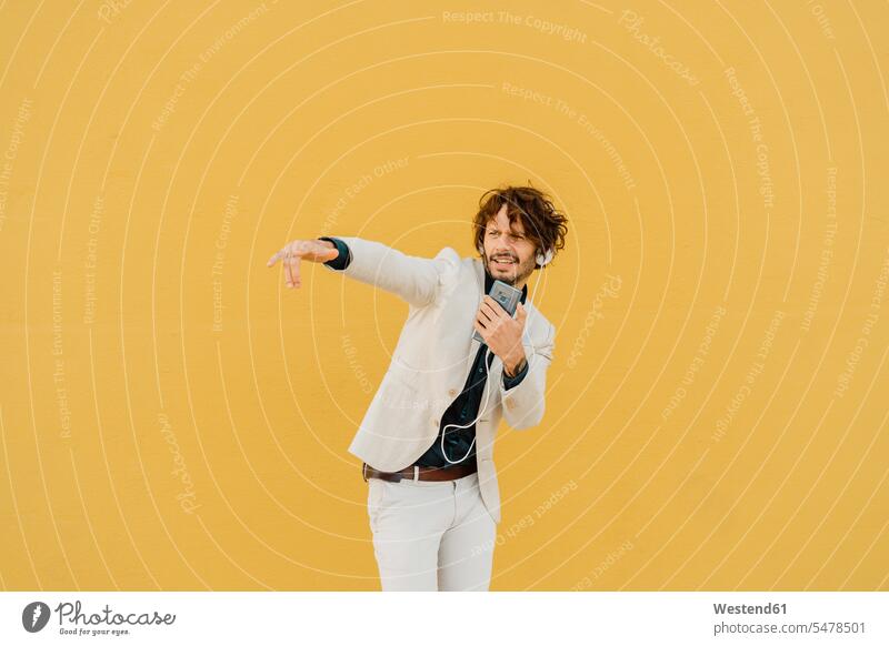 Geschäftsmann singt und tanzt vor der gelben Wand und hört Musik mit Kopfhörern und Smartphone geschäftlich Geschäftsleben Geschäftswelt Geschäftsperson