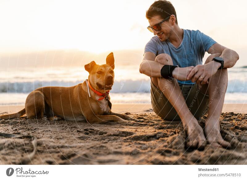 Lächelnder Mann schaut seinen Hund an, während er sich am Strand entspannt Farbaufnahme Farbe Farbfoto Farbphoto Außenaufnahme außen draußen im Freien
