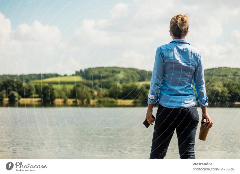 Frau stehend an einem See mit Handy und Kaffee zum Mitnehmen Seen weiblich Frauen steht Mobiltelefon Handies Handys Mobiltelefone Gewässer Wasser Getränk