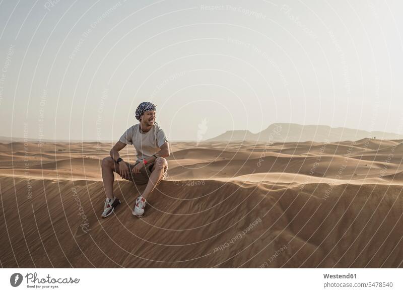 Männlicher Tourist schaut weg, während er auf Sanddünen in der Wüste von Dubai, Vereinigte Arabische Emirate, sitzt Farbaufnahme Farbe Farbfoto Farbphoto