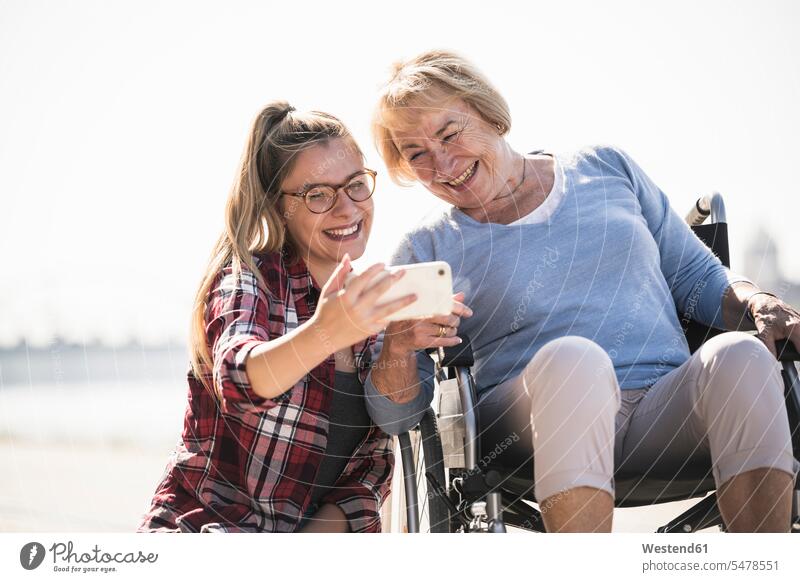 Junge Frau mit ihrer lächelnden Großmutter, die im Rollstuhl sitzt und ein Selfie macht Generation Telekommunikation telefonieren Handies Handys Mobiltelefon