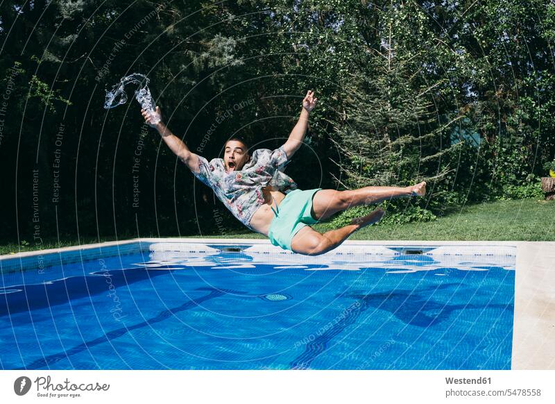 Geschockter junger Mann hält Getränk in der Hand und fällt im Schwimmbad gegen Bäume Farbaufnahme Farbe Farbfoto Farbphoto Spanien Tag Tageslichtaufnahme