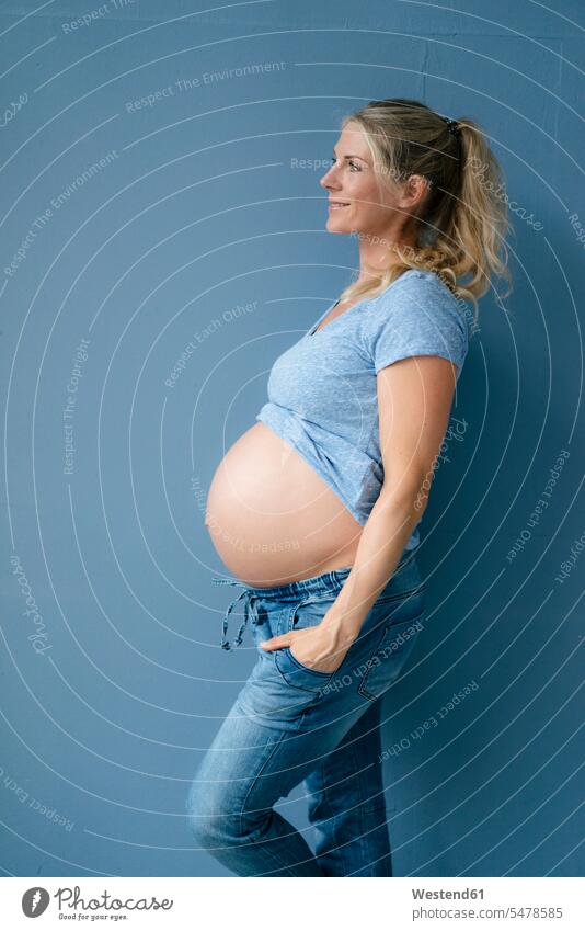 Lächelnde schwangere Frau an blauer Wand stehend Wände Waende steht Schwangere blaues weiblich Frauen Farbe Farbtöne Farbtoene Farbton Farben Erwachsener