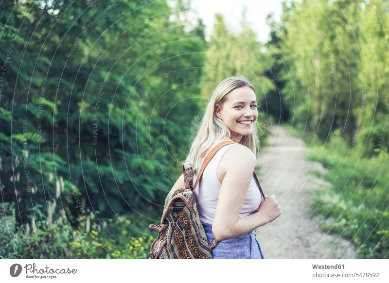 Porträt einer lächelnden jungen Frau mit Rucksack auf einem Waldweg Rucksäcke entspannen relaxen Wanderung Jahreszeiten sommerlich Sommerzeit freuen geniessen