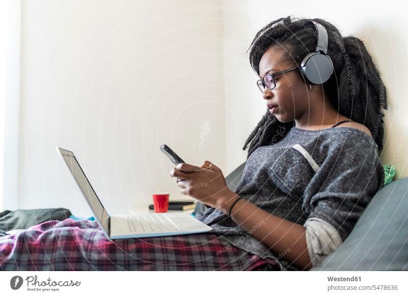 Junge Frau mit Smartphone sitzend mit Laptop und Kopfhörern am Bett zu Hause Leute Menschen People Person Personen 1 Ein ein Mensch eine nur eine Person single
