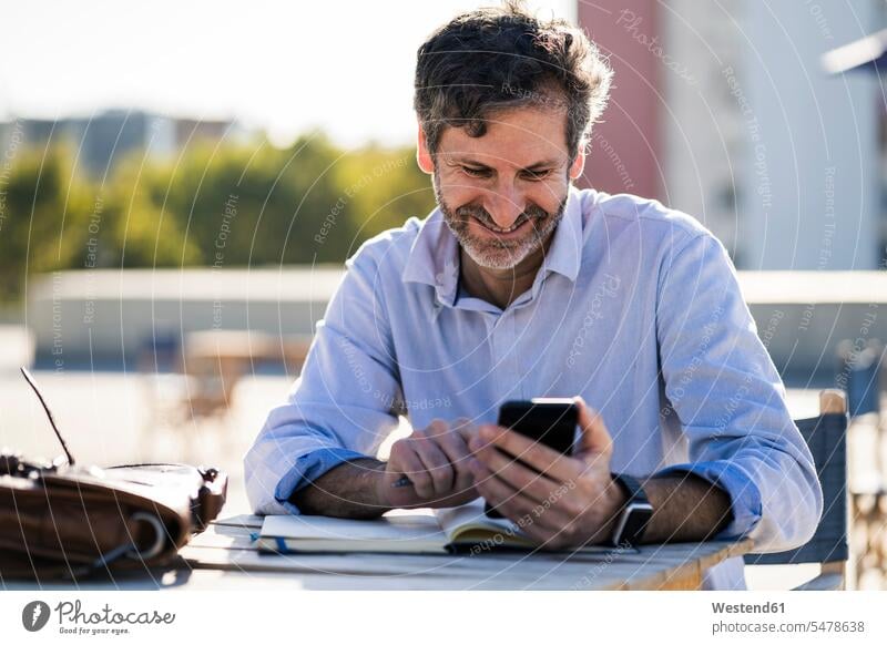 Lächelnde reifen Mann sitzt am Tisch im Freien mit Handy und Notebook lächeln Mobiltelefon Handies Handys Mobiltelefone Männer männlich Notizbuch Notizbücher